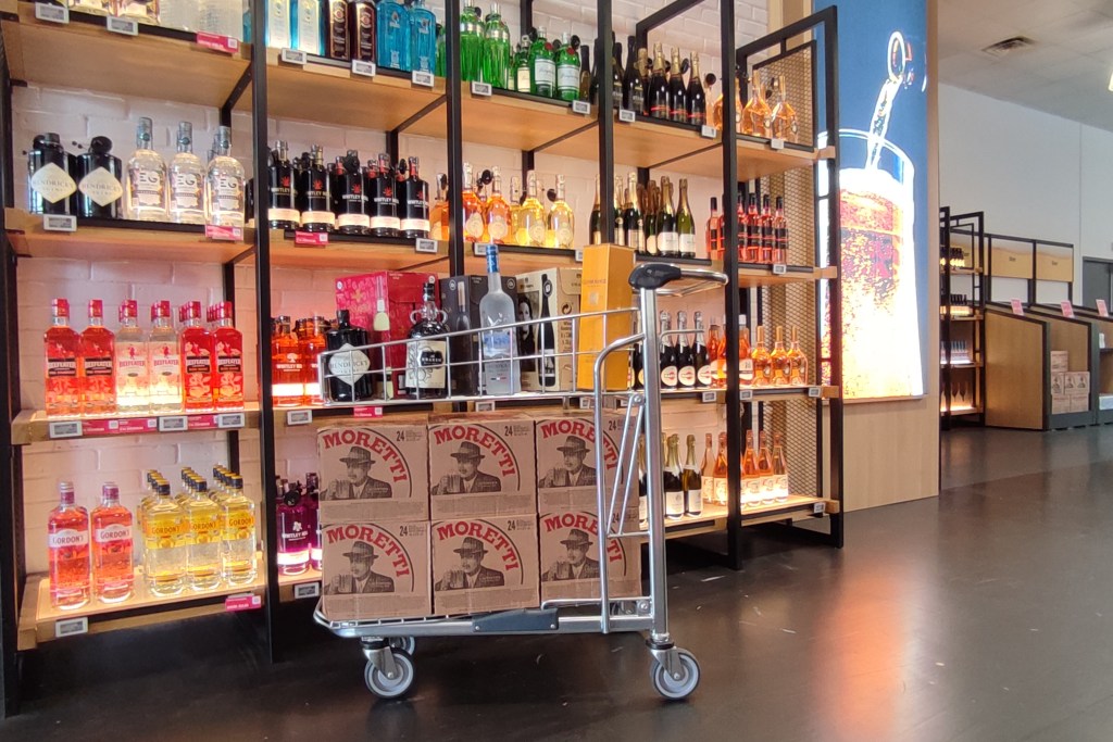 A trolley in the foreground with boxes and bottles of different alcoholic spirits and shelves against a wall lined with spirits and champagne in the background.