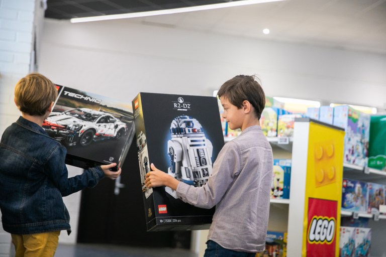 Two children in the Dunkirk duty-free shop, each holding a big, black box - the first with the picture of a car on it and the other R2D2 from Star Wars. The Lego logo shows in the background.