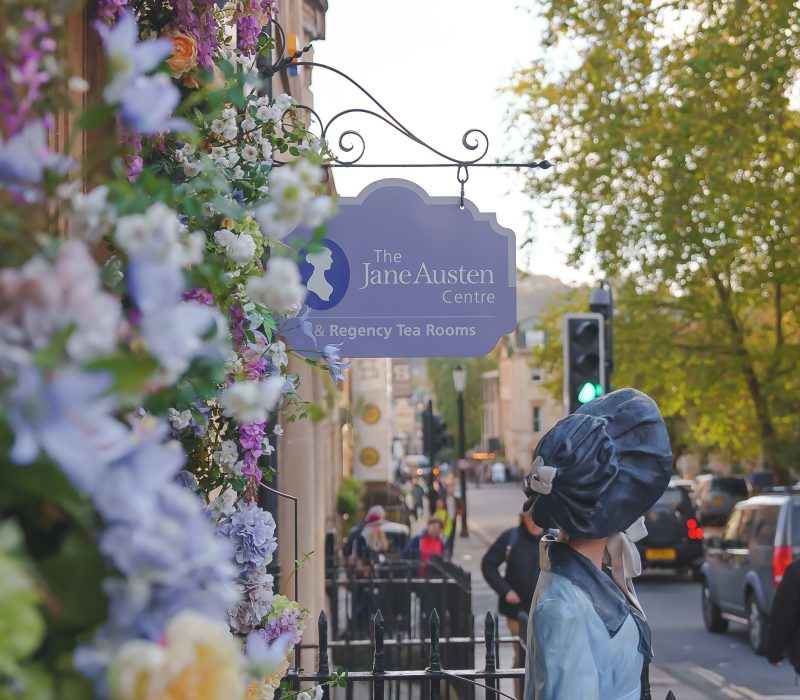 A side shot of the Jane Austen Centre with Jane's statue and sign for the centre present with flowers that decorate the entrance.