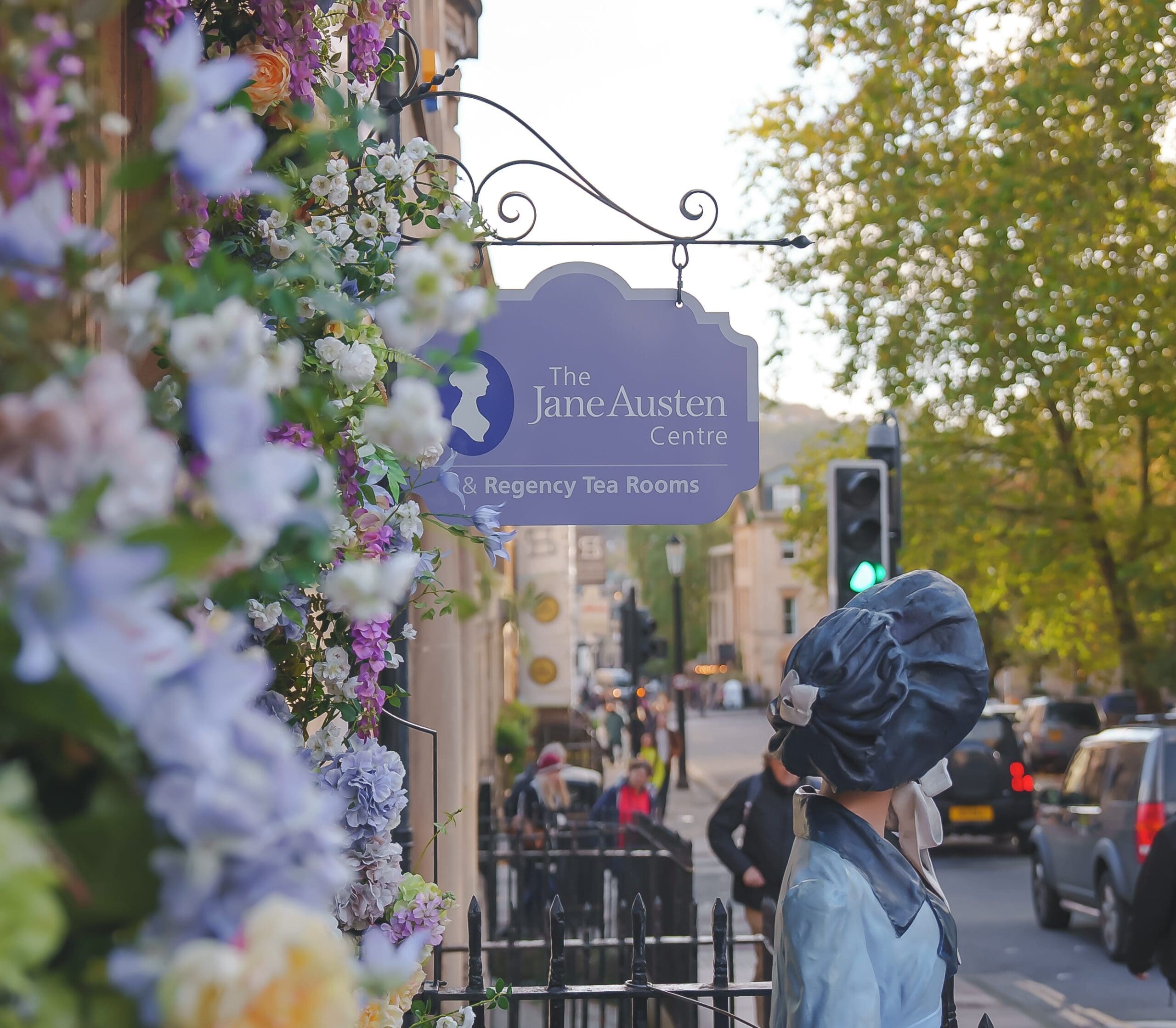 A side shot of the Jane Austen Centre with Jane's statue and sign for the centre present with flowers that decorate the entrance.